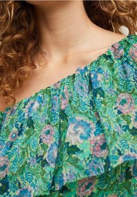 Curly-haired person wearing a green off-shoulder top with a floral pattern in blue, pink, and white tones.