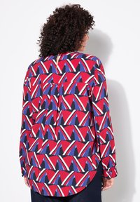 Rode blouse met een geometrisch patroon in blauw, wit en marineblauw. Heeft een afgeronde zoom, lange mouwen en een opening met sleutelhanger aan de achterkant.