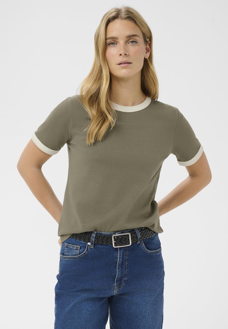 Femeie cu păr blond lung, purtând un tricou verde măsliniu cu margini albe și blugi albaștri, mâinile sprijinite pe șolduri, fundal neutru.