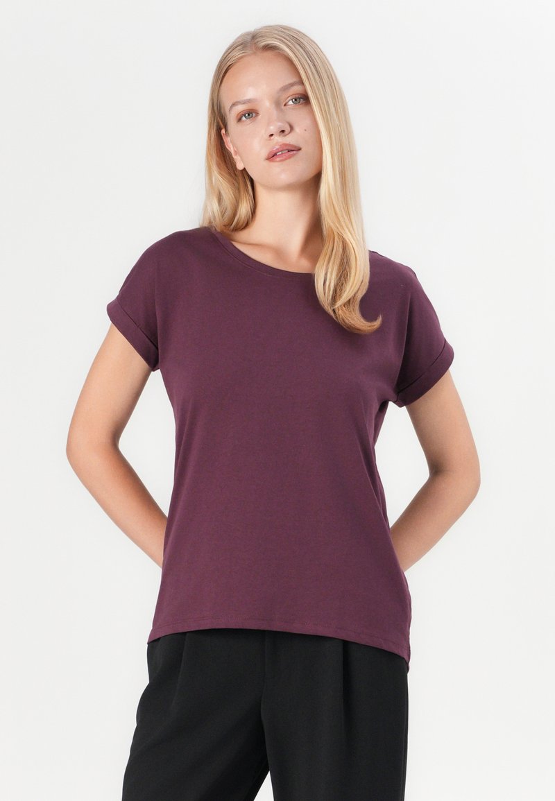 Kurzärmliges T-Shirt mit Rundhalsausschnitt in tiefem Lila, aus weichem Stoff gefertigt. Mit aufgerollten Ärmeln und einem lockeren Schnitt, kombiniert mit schwarzen Hosen.