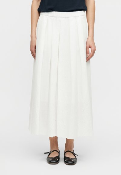 Femme portant une jupe midi plissée blanche avec un ourlet festonné et des chaussures Mary Jane noires à clous argentés, se tenant devant un fond blanc.