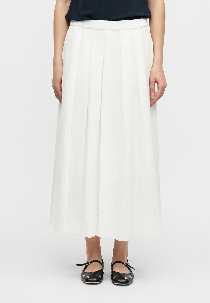 Γυναίκα που φοράει λευκή πλισέ μίν τι φούστα με κυματιστό τελείωμα και μαύρα παπούτσια Mary Jane με ασημένιες καρφίτσες, στέκεται μπροστά σε λευκό φόντο.