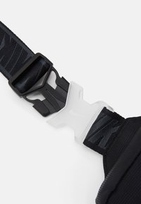 Svart nylonrem med justerbar svart spänne och genomskinlig plastfäste med subtilt präglad logotyp på vit bakgrund.