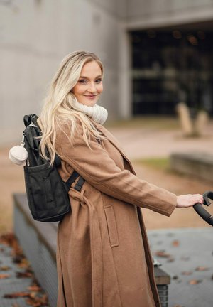 Schwarzer Rucksack mit mehreren Taschen, Stoffstruktur und einem beigefarbenen, runden Schlüsselanhänger, getragen über einem braunen Mantel.