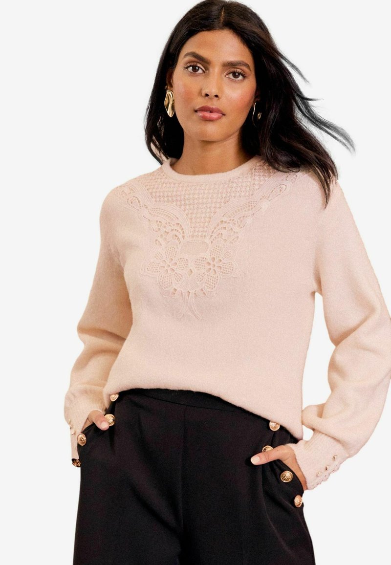 Gaiša rozā džemperis ar ziedu mežģīņu detalizāciju priekšpusē, apaļa apkakle un garas piedurknes. Pogas pie aprocēm piešķir rotājumu.