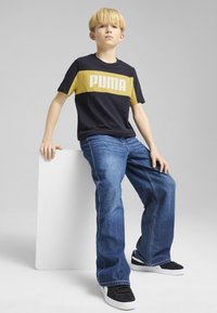 Marineblauwe en gele t-shirt met korte mouwen en het "PUMA" logo, gecombineerd met blauwe spijkerbroeken en zwarte sneakers, zittend tegen een witte blok.