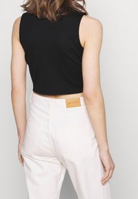 Žena v černém bezrukávovém crop topu a bílých jeanech s vysokým pasem, na zadním pásku s logem značky Monki.