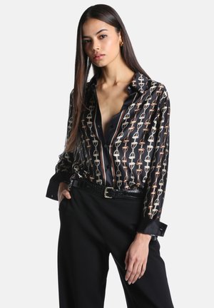 Femme aux longs cheveux raides portant un chemisier en satin noir à motifs, rentré dans un pantalon noir taille haute avec une ceinture noire.