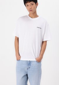 Camiseta blanca de manga corta hecha de tela suave, con una pequeña impresión negra que dice "Club 1990" en la parte izquierda del pecho, combinada con pantalones vaqueros de un azul claro.