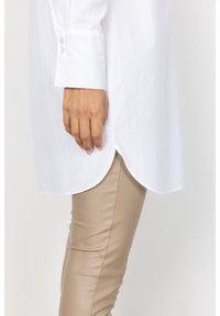 Camicia bianca a maniche lunghe con orlo curvo e polsini abbottonati, abbinata a pantaloni aderenti beige, che mostrano un tessuto liscio e un design su misura.