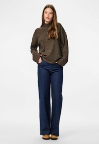 Une femme se tient debout les mains dans les poches, portant un pull côtelé marron, un jean large bleu et des chaussures beiges sur un fond blanc.