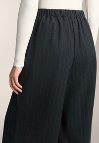 Črne široke hlače z ozkimi črtami in elastičnim pasom. Tkanina deluje teksturirano in gladko, z rahlim leskom črt.