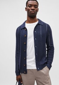 Sötétkék kötött kardigán, gallérral, gombos záródással és függőleges nyitott varrási mintákkal. Fehér ing fölött viselve.