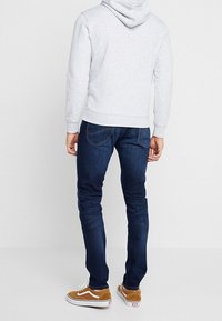 Personne portant un sweat à capuche gris clair, un jean slim bleu foncé et des baskets marron, debout de dos sur un fond uni.