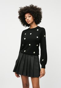 Suéter negro con lunares dorados, con mangas largas y cuello redondo. Combinado con una falda negra plisada, que destaca la textura y la forma.