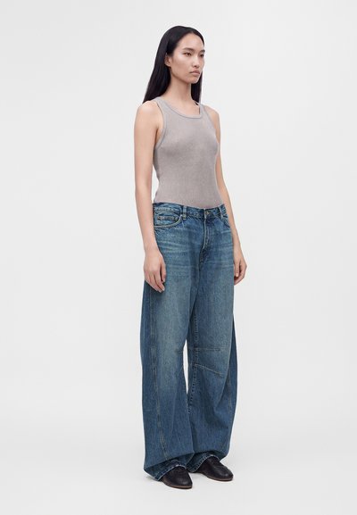 Joven mujer de pie, con una camiseta gris sin mangas, vaqueros azules de pierna ancha y zapatos negros, frente a un fondo claro y liso.