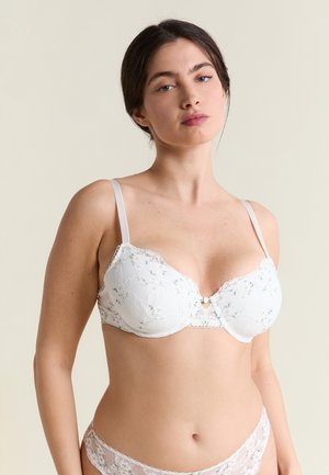 Donna con capelli scuri che indossa un reggiseno in pizzo floreale bianco e mutandine abbinate contro uno sfondo beige uniforme.
