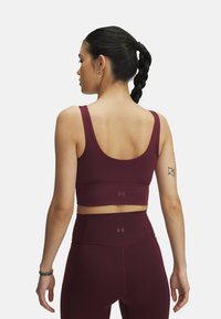 Burgundy athletic crop top with wide straps, smooth texture, and logo detail on the back, paired with high-waisted leggings.