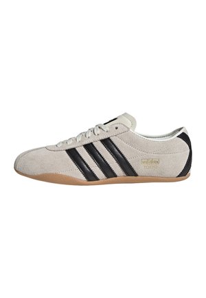 Lichtbeige suède sneaker met drie zwarte strepen, rubberen zool, witte veters en gouden ""adidas TOKYO""-opschrift aan de zijkant.