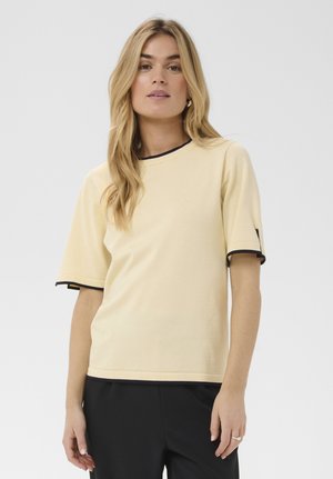 Kvinde med langt blond hår iført en beige kortærmet top med sort kant og sorte bukser, står foran en neutral baggrund.