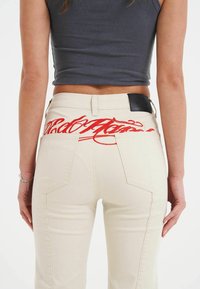Beiga denimjeans med kontrasterande röd broderad text på bakfickan, i kombination med en åtsittande grå cropped top.