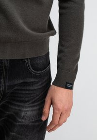 Dunkelgrüner Strickpullover mit gerippten Bündchen, kombiniert mit dunklen Jeans, die eine dezente Verwaschung und ein kontrastierendes Nähdetail aufweisen.