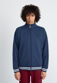 Navy zip-up jacket with high collar, white striped cuffs and hem, textured fabric, featuring a small logo on the chest.