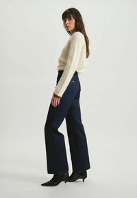 Pull en tricot de couleur crème clair, jean en denim foncé taille haute évasé et bottines noires à bout pointu, se tenant devant un fond neutre.