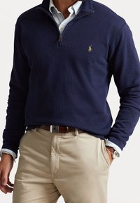 Sudadera de media cremallera en color marino, hecha de un tejido suave, con una textura acanalada y un pequeño logotipo bordado. Lleva puesta sobre una camisa de cuello azul claro.