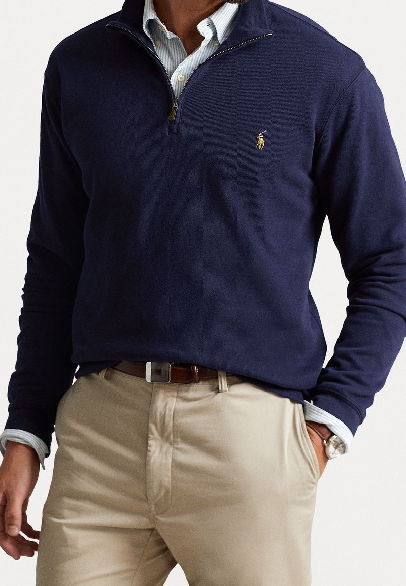 Sudadera de media cremallera en color marino, hecha de un tejido suave, con una textura acanalada y un pequeño logotipo bordado. Lleva puesta sobre una camisa de cuello azul claro.