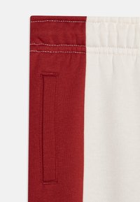 I pantaloni della tuta rossi e crema presentano un elastico in vita con cuciture visibili e una tasca laterale nella parte rossa. Texture del tessuto morbida.