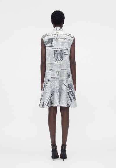 Άτομο που φοράει αμάνικο φόρεμα με τύπωμα εφημερίδας, όρθιο με την πλάτη προς τα εμπρός, συνδυασμένο με μαύρα παπούτσια με ψηλό τακούνι.