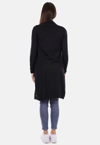 Cardigan lungo nero con colletto, texture morbida, spacchi laterali e dettagli con bottoni all'orlo, indossato sopra jeans blu chiaro e sneaker bianche.