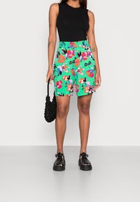 Shorts verts à fleurs avec une taille haute et une ceinture élastique, assortis à un haut sans manches noir et des sandales noires à plateforme, portant un sac noir texturé.