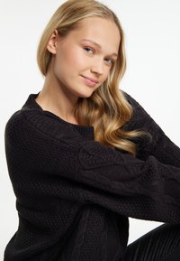 Schwarzer Strickpullover mit einem strukturierten Muster und Zopfmuster. Das Material wirkt weich mit einem lockeren Schnitt und runder Ausschnitt.