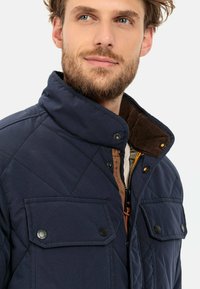 Marineblau gesteppte Jacke mit hohem Kragen, ausgestattet mit zwei aufgesetzten Fronttaschen, braunem Cord-Lining und goldfarbenen Hardware-Details.