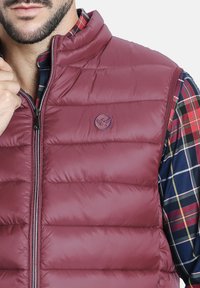 Chaleco puffer burdeos con costuras horizontales, cierre de cremallera y detalle del logo. Superpuesto sobre una camisa de cuadros en colores oscuros y brillantes.