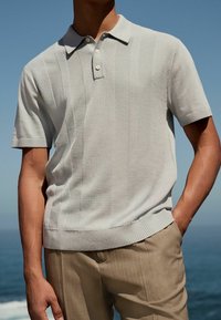 Lichtgrijze polo met korte mouwen, voorzien van een gestructureerd gestreept patroon, drie knopen bij de hals en ribgebreide kraag en zoom. Gecombineerd met beige broek.