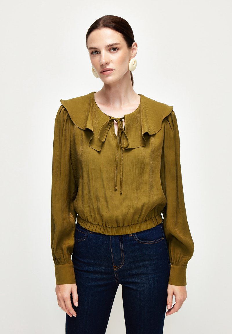 Olivengrøn bluse med rynket krave og bindebånd ved halsen, lange ærmer og elastisk talje, parret med mørkeblå jeans.