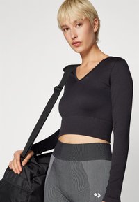 Black ribbed crop top with long sleeves and a V-neckline, paired with gray patterned leggings. Model holds a black gym bag.