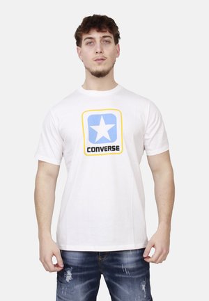 Giovane uomo che indossa una t-shirt bianca con un logo a stella blu e testo "CONVERSE", abbinata a jeans blu strappati, in piedi su uno sfondo bianco.