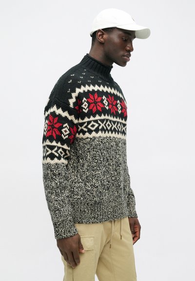 Crno-bijeli pleteni pulover s crvenim cvjetnim uzorcima, visoki ovratnik i strukturirani tijelo. Model nosi bež cargo hlače i bijelu kapu.