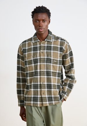 Camisa de cuadros marrón y verde con acentos blancos, que cuenta con un cuello tipo botón y un detalle de bolsillo delantero, hecha de tejido texturizado.