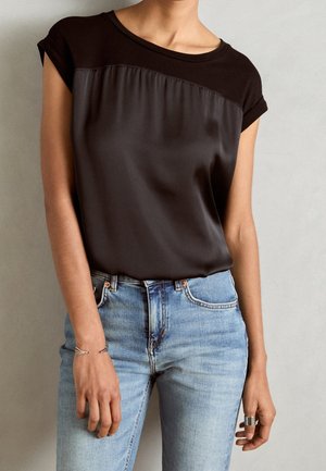 Personne portant un haut noir à manches courtes avec un panneau soyeux et un jean taille haute bleu clair, debout devant un fond clair uni.