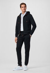 Zwarte hoodie met oranje accenten, ritszakken en joggers. Gecombineerd met witte sneakers, gedragen over een witte poloshirt.