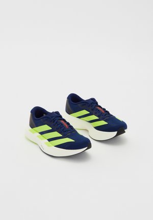 Marineblauwe hardloopschoenen met neon groene strepen, gestructureerde bovenkant, gewatteerde witte zool, vetersluiting en rode accenten op de tong.