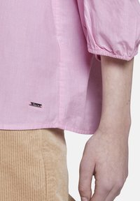 Blusa rosa, leggera, a maniche lunghe con polsini arricciati e una piccola etichetta con logo vicino all'orlo. Il tessuto appare morbido con una sottile trama.