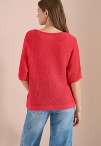Femme aux cheveux bruns et raides porte un pull rouge ample à manches courtes et un jean bleu clair, debout face à un mur beige.