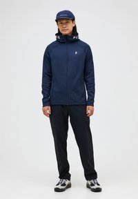 Chaqueta de punto azul marino con cuello alto, texto "PAZ" en blanco y logotipo en contraste; combinada con pantalones negros y zapatos de senderismo grises.