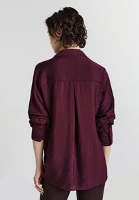 Donna con capelli corti e ricci che indossa una camicia a maniche lunghe, arrotolata, di tessuto texture di un colore viola scuro, vista da dietro su uno sfondo semplice.